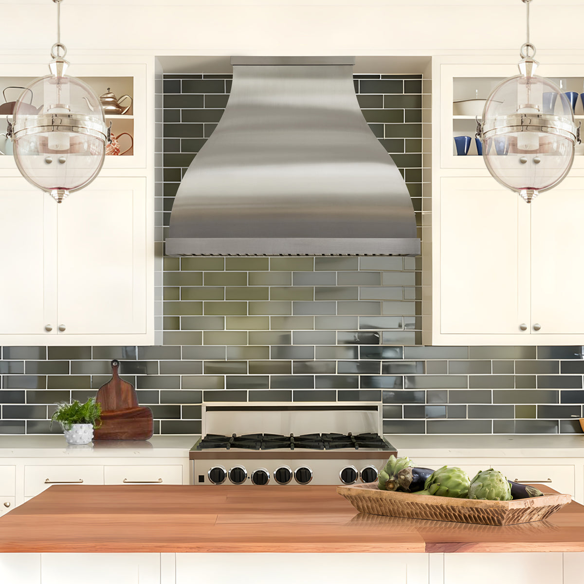 a modern stainless steel kitchen range hood mounted above a gas stove in a bright, modern kitchen with white cabinets, a green subway tile backsplash, and glass pendant lights